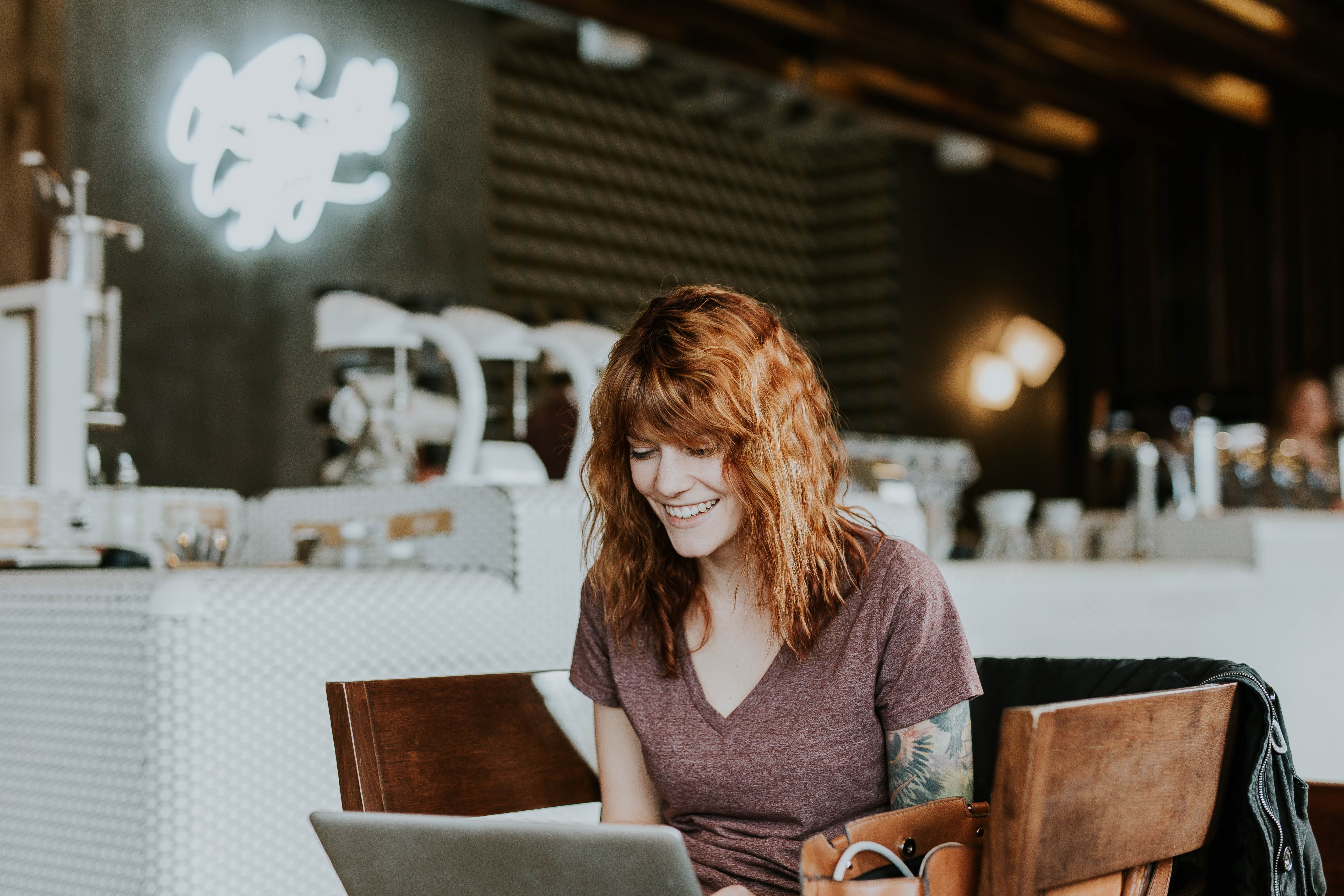 A woman with red hair works on a laptop in a space that looks like a coffee shop
